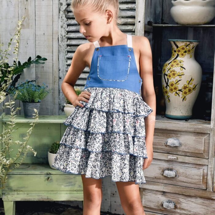 Girl's summer dungaree dress with blue denim cotton top and navy, red, white floral ruffle bottom with adjustable cross back straps. Model dungarees dress with ruffles for girls, girls of the brand of children's fashion LA. FAUTE A VOLTAIRE