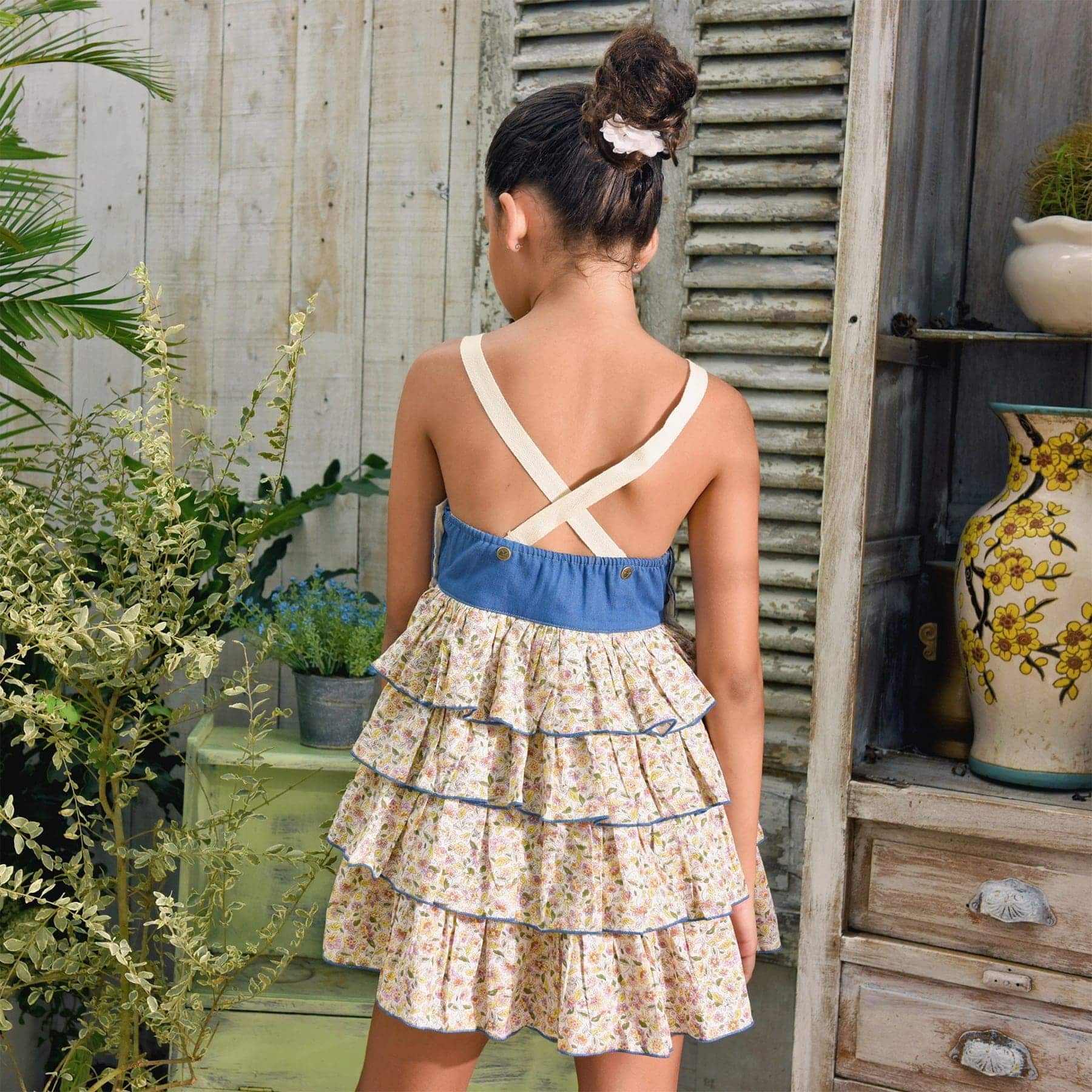 Girl's summer dungaree dress with blue denim cotton top and yellow and light blue floral ruffle bottom, adjustable cross-back straps. Model dungarees dress with ruffles for girls, girls of the brand of children's fashion LA. FAUTE A VOLTAIRE