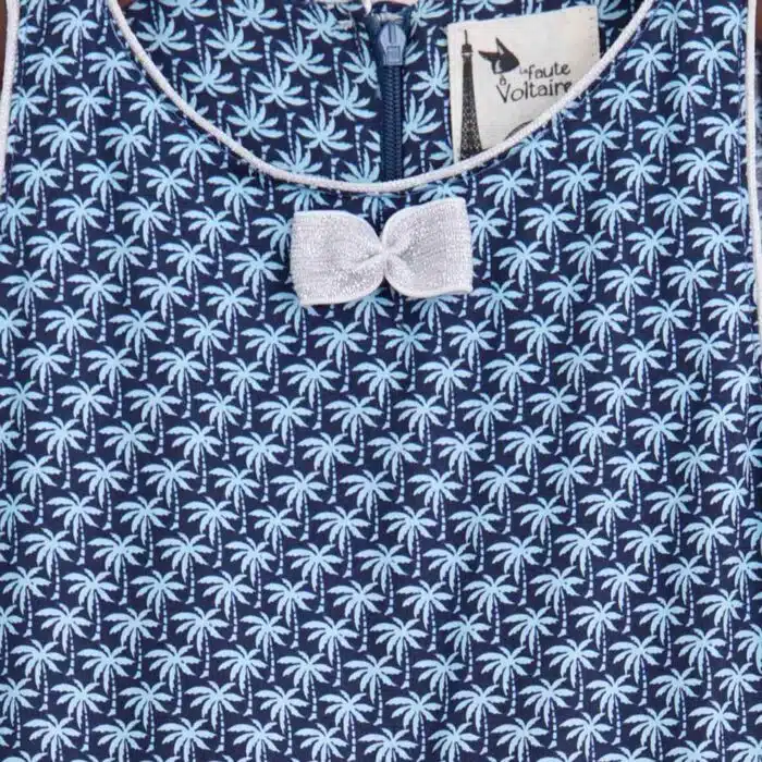 Belle robe qui tourne pour filles de 2 à 16 ans en coton bleu marine imprimé de palmier en bleu clair. Une ceinture à rayures multicolore à paillettes et noeud argenté sur la poitrine, modèle HEPBURN de la marque de mode pour enfants LA FAUTE A VOLTAIRE