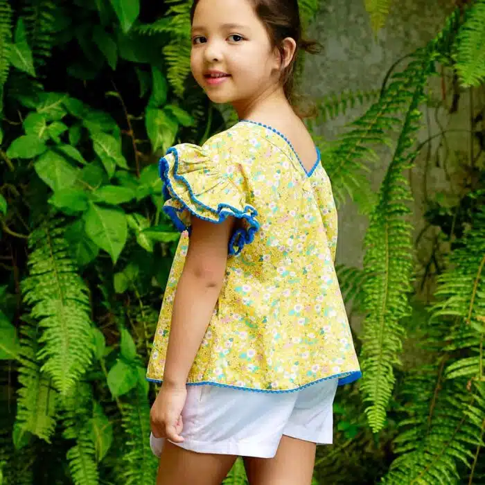 Jolie blouse fleurie ample, à manches papillon double rangée de volants en coton fleurs liberty jaune, bleu, rose pâle. Originale avec sa double rangée de volants bordée de pompons bleu roi. Col V dans la nuque souligné de pompons bleu, bas de la blouse souligné de pompons bleu roi. Blouse été pour filles, fillettes et ados de 2 à 16 ans, de la marque de mode pour enfants LA FAUTE A VOLTAIRE