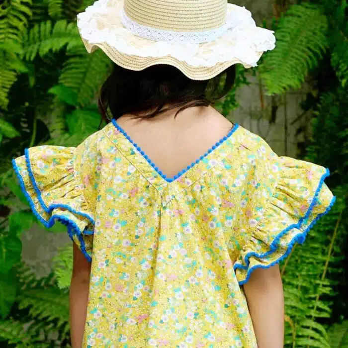 Jolie robe fleurie liberty jaune à petites fleurs rose, bleu turquoise, vert pâle, avec une double rangée de volants aux manches style papillon, col V dans la nuque, pompons bleu roi autour du col, des manches et. bas de la robe. Robe été pour filles, fillettes et ados de la marque de mode La Faute à Voltaire