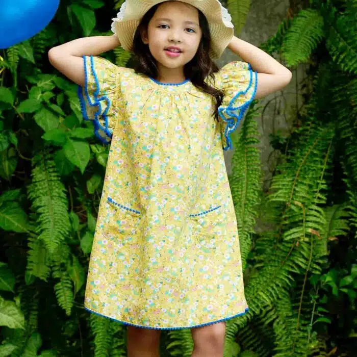 Jolie robe fleurie liberty jaune à petites fleurs rose, bleu turquoise, vert pâle, avec une double rangée de volants aux manches style papillon, col V dans la nuque, pompons bleu roi autour du col, des manches et. bas de la robe. Robe été pour filles, fillettes et ados de la marque de mode La Faute à Voltaire