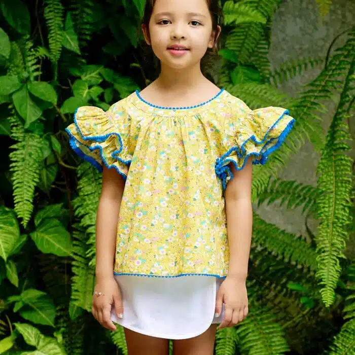 Jolie blouse fleurie ample, à manches papillon double rangée de volants en coton fleurs liberty jaune, bleu, rose pâle. Originale avec sa double rangée de volants bordée de pompons bleu roi. Col V dans la nuque souligné de pompons bleu, bas de la blouse souligné de pompons bleu roi. Blouse été pour filles, fillettes et ados de 2 à 16 ans, de la marque de mode pour enfants LA FAUTE A VOLTAIRE