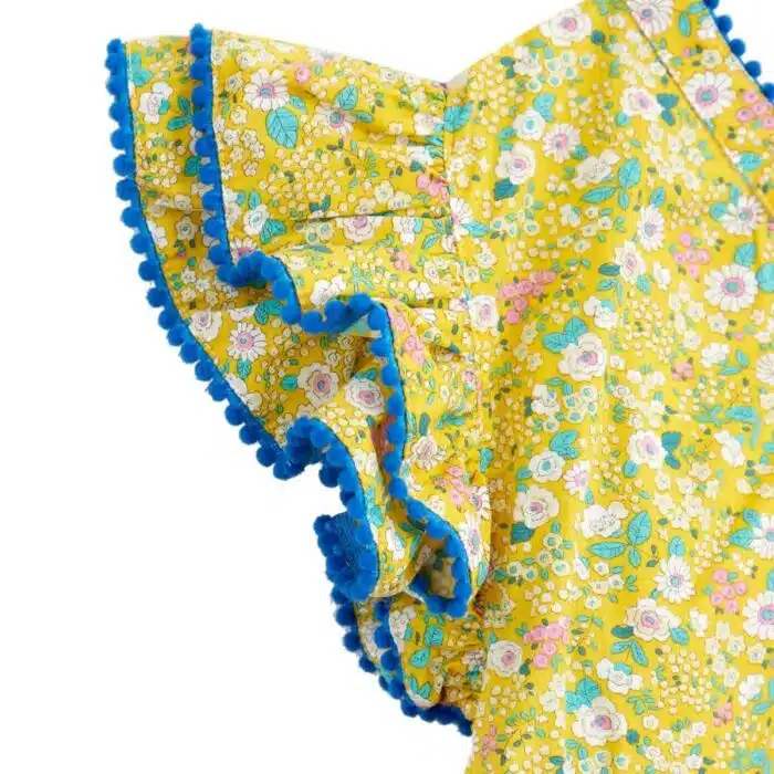 Jolie robe fleurie liberty jaune à petites fleurs rose, bleu turquoise, vert pâle, avec une double rangée de volants aux manches style papillon, col V dans la nuque, pompons bleu roi autour du col, des manches et. bas de la robe. Robe été pour filles, fillettes et ados de la marque de mode La Faute à Voltaire