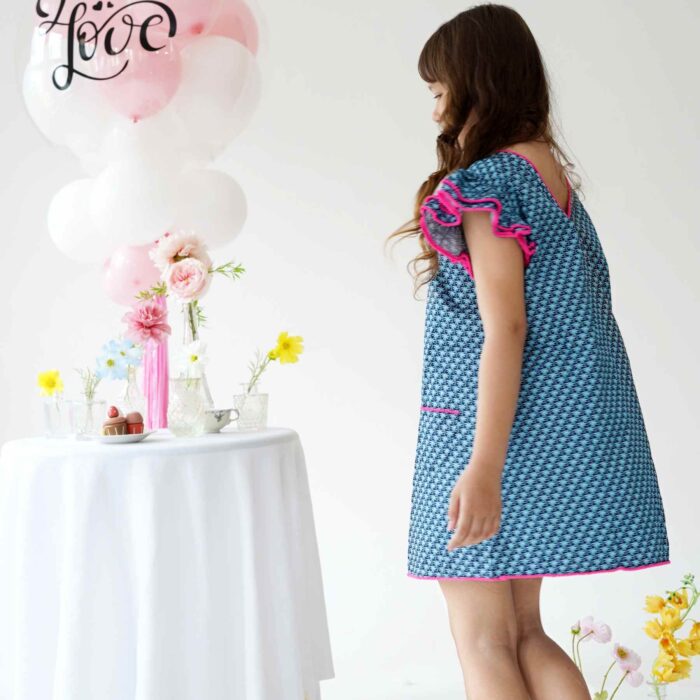 Une Magnifique Robe d'Été Bleu Foncé, Manches à Volants bordées de Pompons Rose Fuchsia, Col V dans le Nuque de la Marque de Mode Française pour Enfants La Faute à Voltaire.