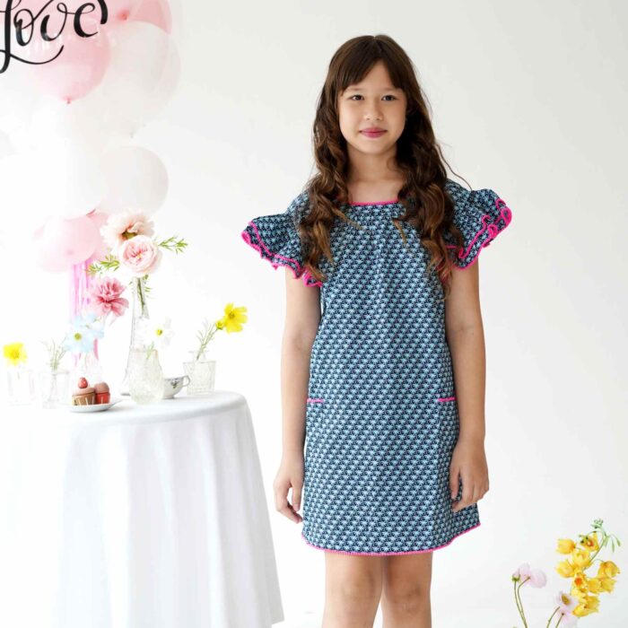 Une Magnifique Robe d'Été Bleu Foncé, Manches à Volants bordées de Pompons Rose Fuchsia, Col V dans le Nuque de la Marque de Mode Française pour Enfants La Faute à Voltaire.