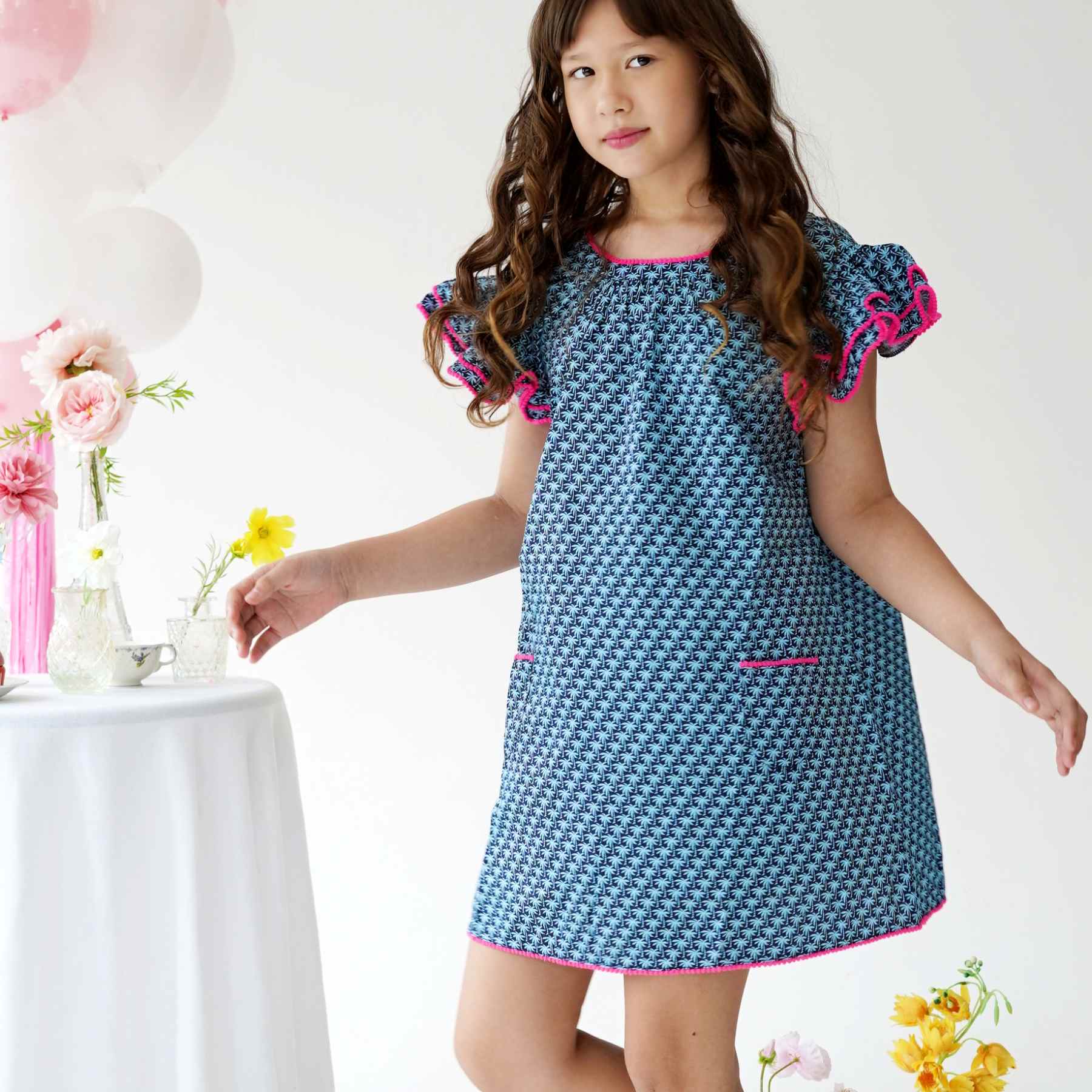 Une Magnifique Robe d'Été Bleu Foncé, Manches à Volants bordées de Pompons Rose Fuchsia, Col V dans le Nuque de la Marque de Mode Française pour Enfants La Faute à Voltaire.