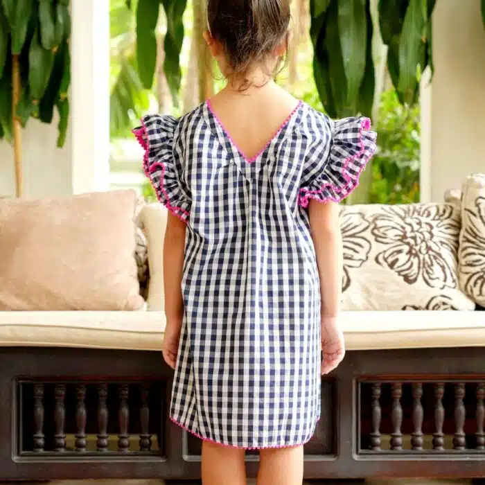 Jolie robe à carreaux vichy bleu marine et blanc, à manches papillons à volants, col rond devant et col V dans la nuque, rangée de pompons rose fuchsia autour des volants, col et bas de la robe. Robe été pour filles, fillettes et ados de la marque de mode pour enfants La Faute à Voltaire