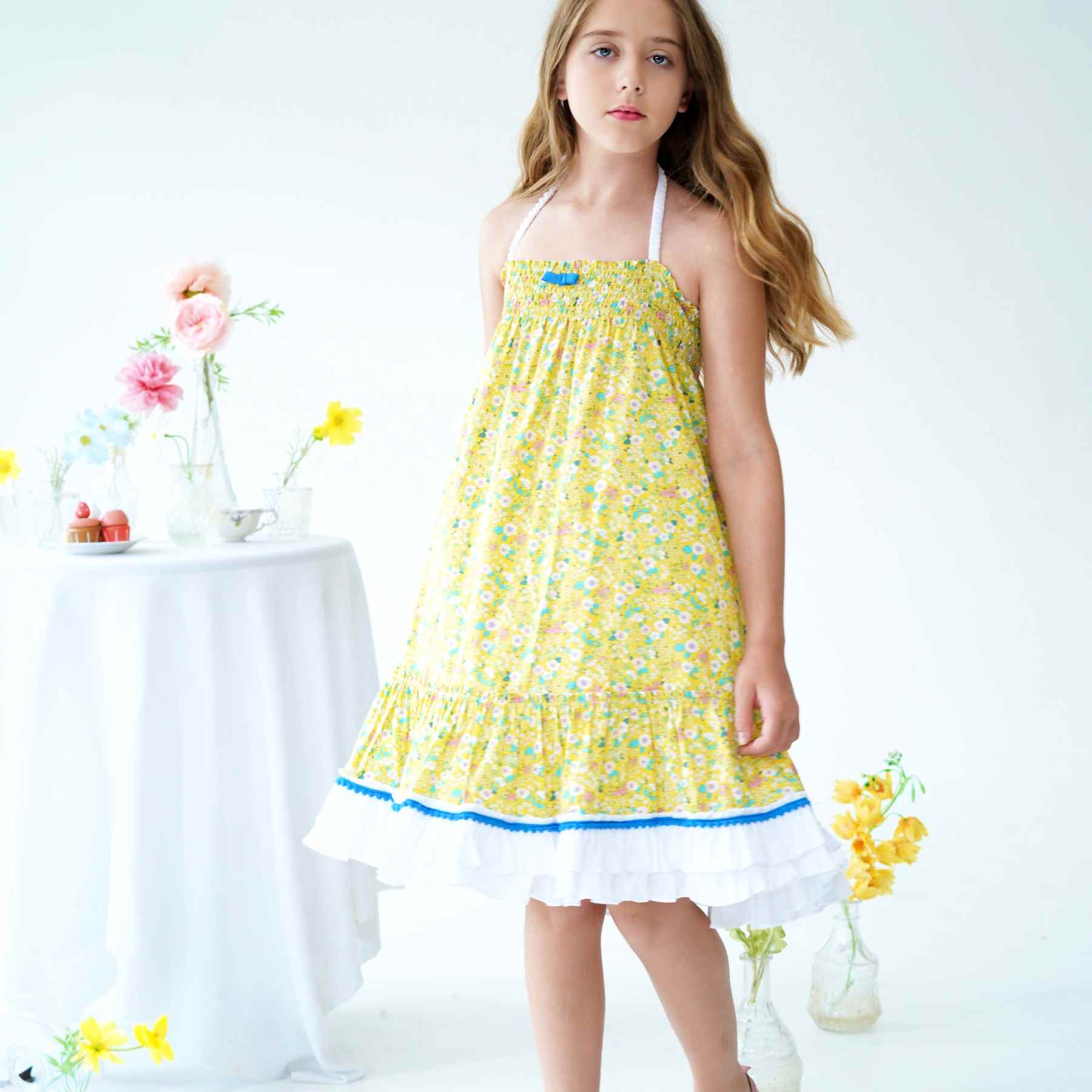 Robe fille fleurie liberty jaune, longueur midi, col smocks, volants blancs en bas de la robe bordés de pompons bleu roi. É-en-1, cette robe se transforme en jupe longue bohème. Tailles disponibles du 2 au 16 ans.