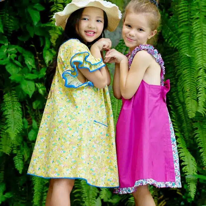 Jolie robe fleurie liberty jaune à petites fleurs liberty rose, bleu turquoise, vert pâle, avec une double rangée de volants aux manches style papillon, col V dans la nuque, pompons bleu roi autour du col, des manches et. bas de la robe. Robe été pour filles, fillettes et ados de la marque de mode La Faute à Voltaire