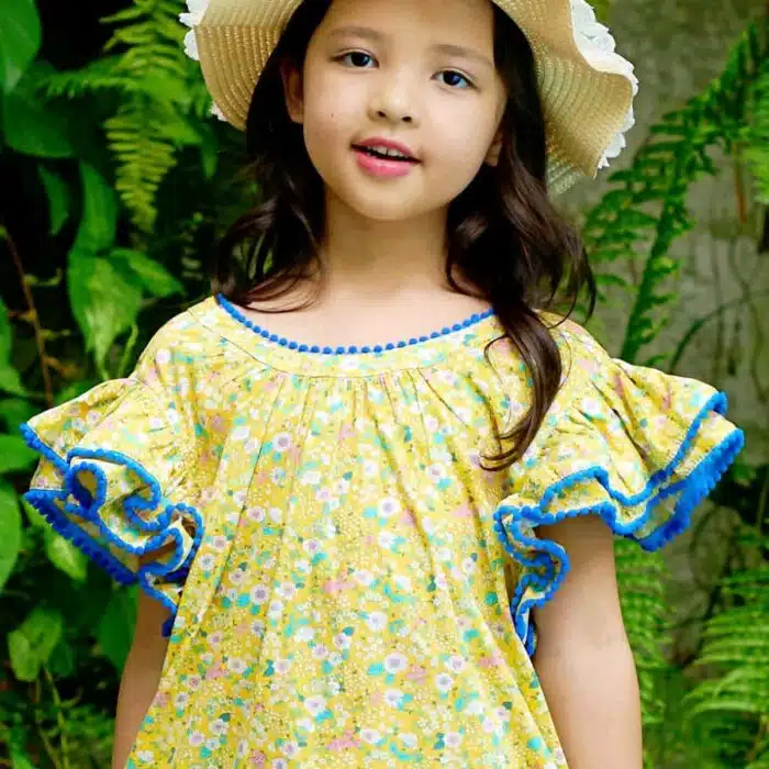 Jolie robe fleurie liberty jaune à petites fleurs rose, bleu turquoise, vert pâle, avec une double rangée de volants aux manches style papillon, col V dans la nuque, pompons bleu roi autour du col, des manches et. bas de la robe. Robe été pour filles, fillettes et ados de la marque de mode La Faute à Voltaire