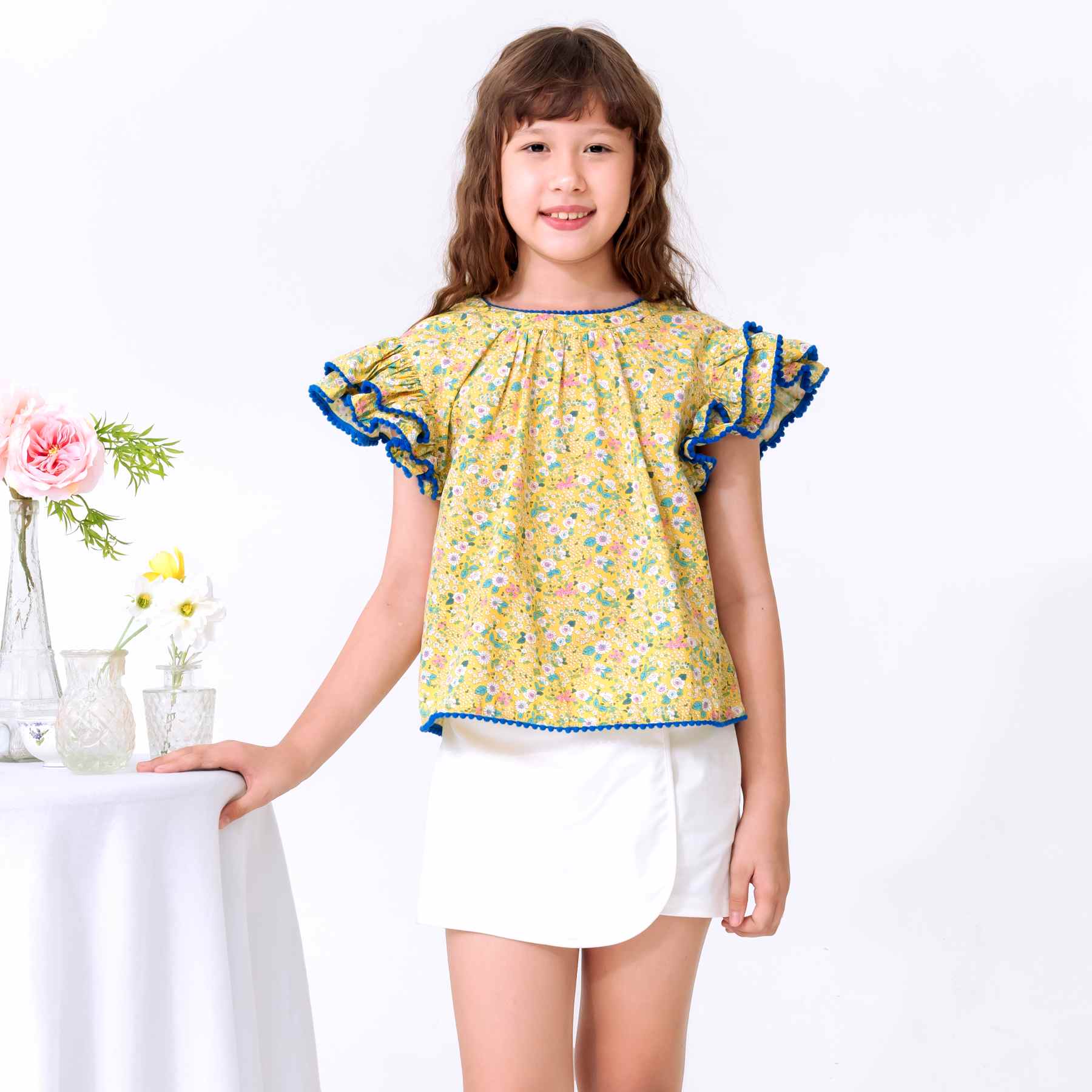 Une Adorable Blouse Été pour Filles, Fillettes et Ados en Coton Fleuri Jaune Pastel, Bleu, Mauve avec Manches Courtes Double Rangées de Volants Bordés de Pompons Courts Bleu Roi, Col V dans la Nuque de la Marque de Mode Française pour Enfants La Faute à Voltaire
