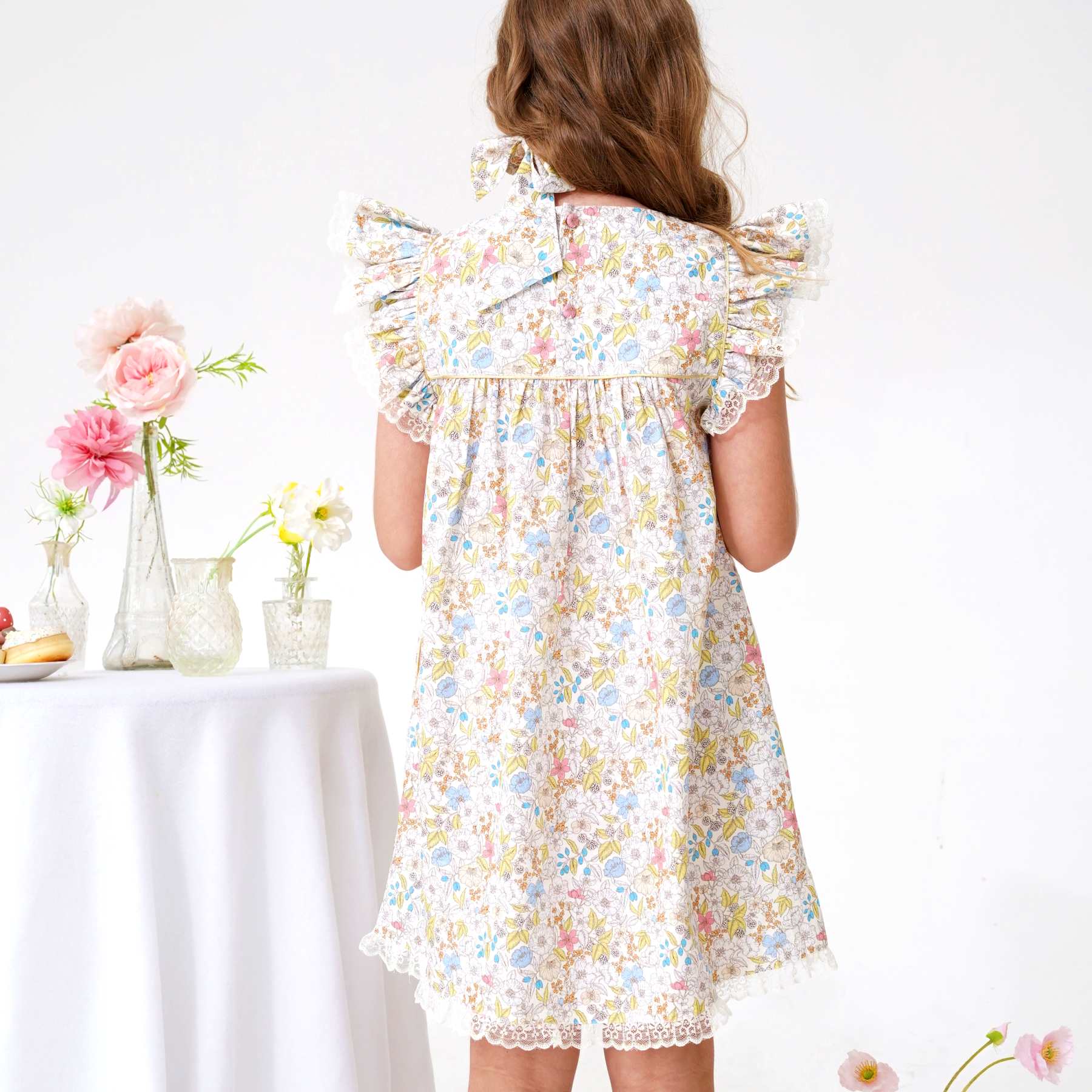 Photo portée du dos de la robe de cérémonie fille fleurie liberty jaune, rose, bleu ciel avec manches à volants bordée de fine dentelle beige mordorée rappelée en bas de la robe, col rond avec fermeture dans la nuque avec petits boutons ronds, biais doré sur le plastron et les emmanchures. Robe idéale pour demoiselle d'honneur et tous les évènements chics de l'été. Disponible du 2 au 16 ans.