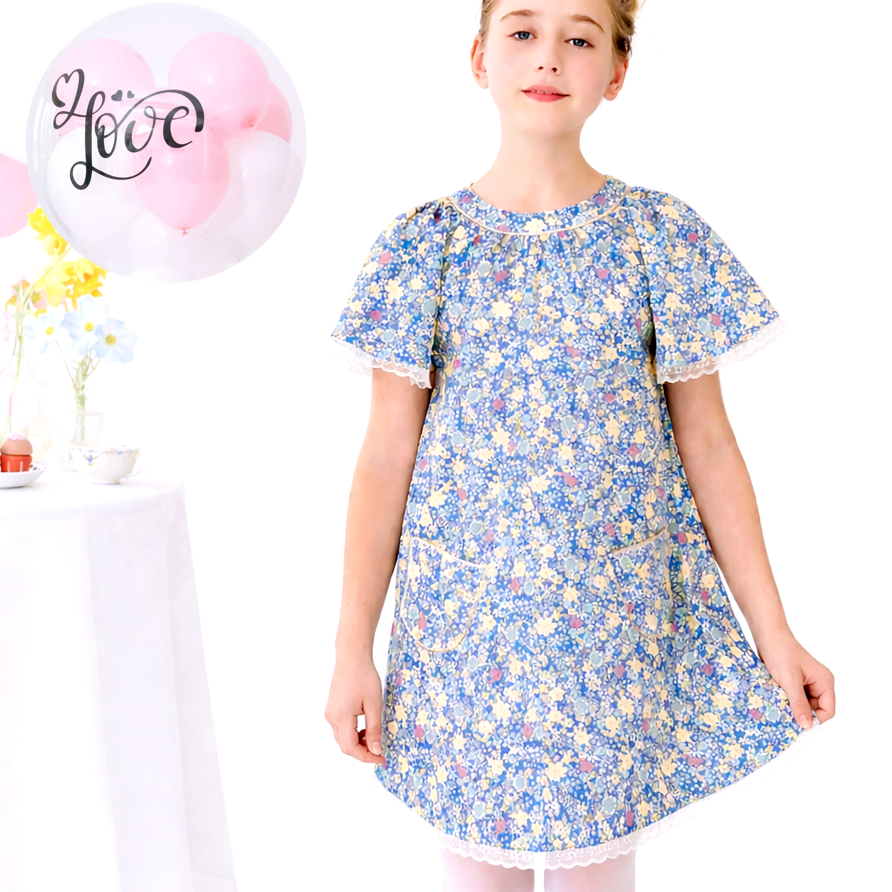 Photo portée de la robe de cérémonie en coton liberty bleu foncé et jaune pour fille. Encolure ronde bordée d'un biais doré. Manches papillon courtes bordées de file dentelle beige mordorée, bas de la robe niveau genou avec dentelle assortie aux manches. Modèle de robe coupe A ample "Butterfly" pour filles disponible de la taille 2 ans à la taille 16 ans de la marque de mode Française pour enfant La Faute à Voltaire.