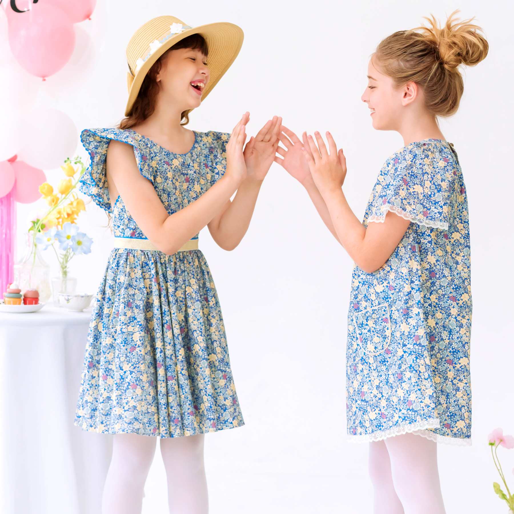 Photo portée de deux robes de cérémonie pour filles en coton fleuri liberty bleu foncé et jaune. L'une à la coupe patineuse avec bretelles à volants appelée Angel et l'autre à manches papillon dans le même tissu, appelé Butterfly.