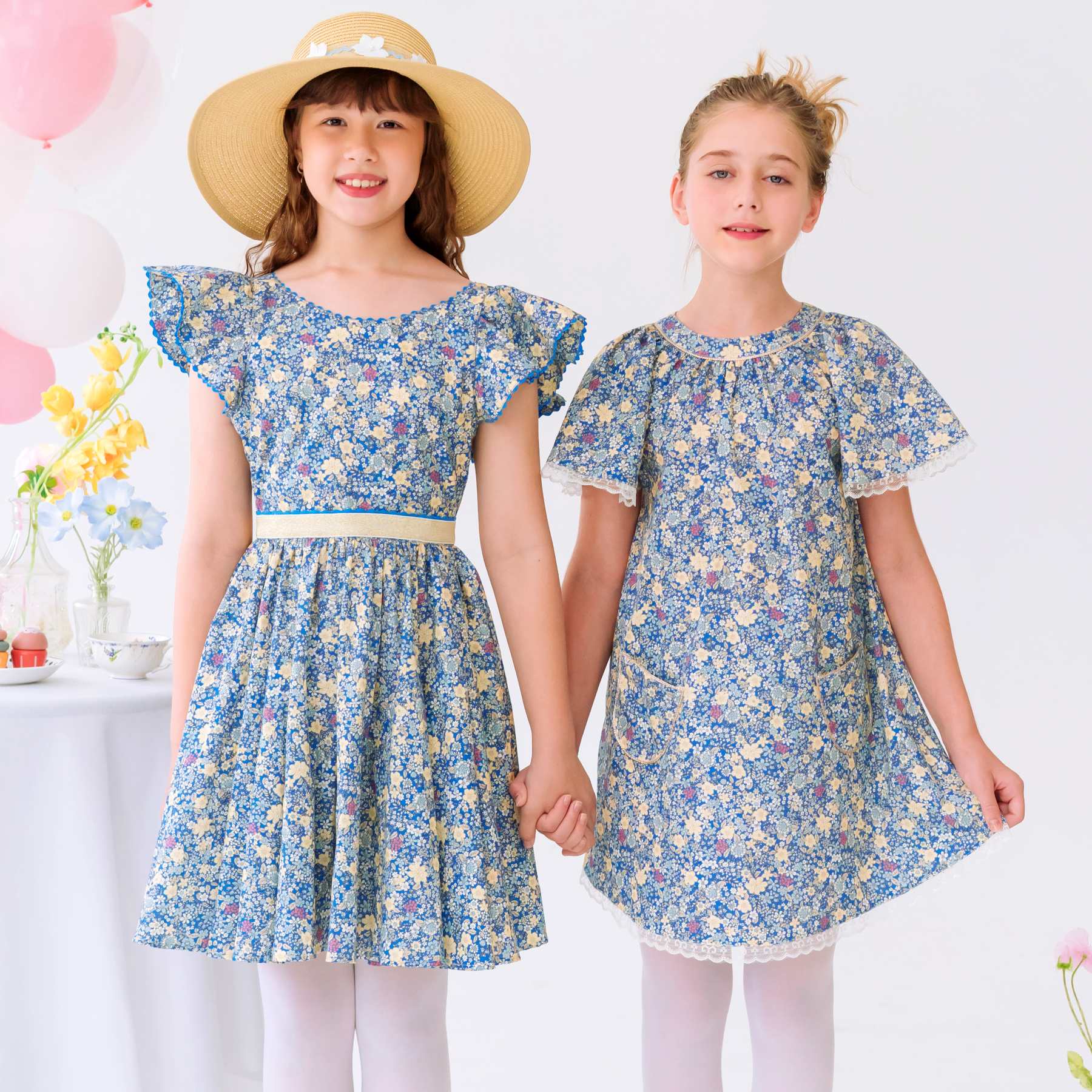 Photo portée de deux robes de cérémonie pour filles en coton fleuri liberty bleu foncé et jaune. L'une à la coupe patineuse avec bretelles à volants appelée Angel et l'autre à manches papillon dans le même tissu, appelé Butterfly.