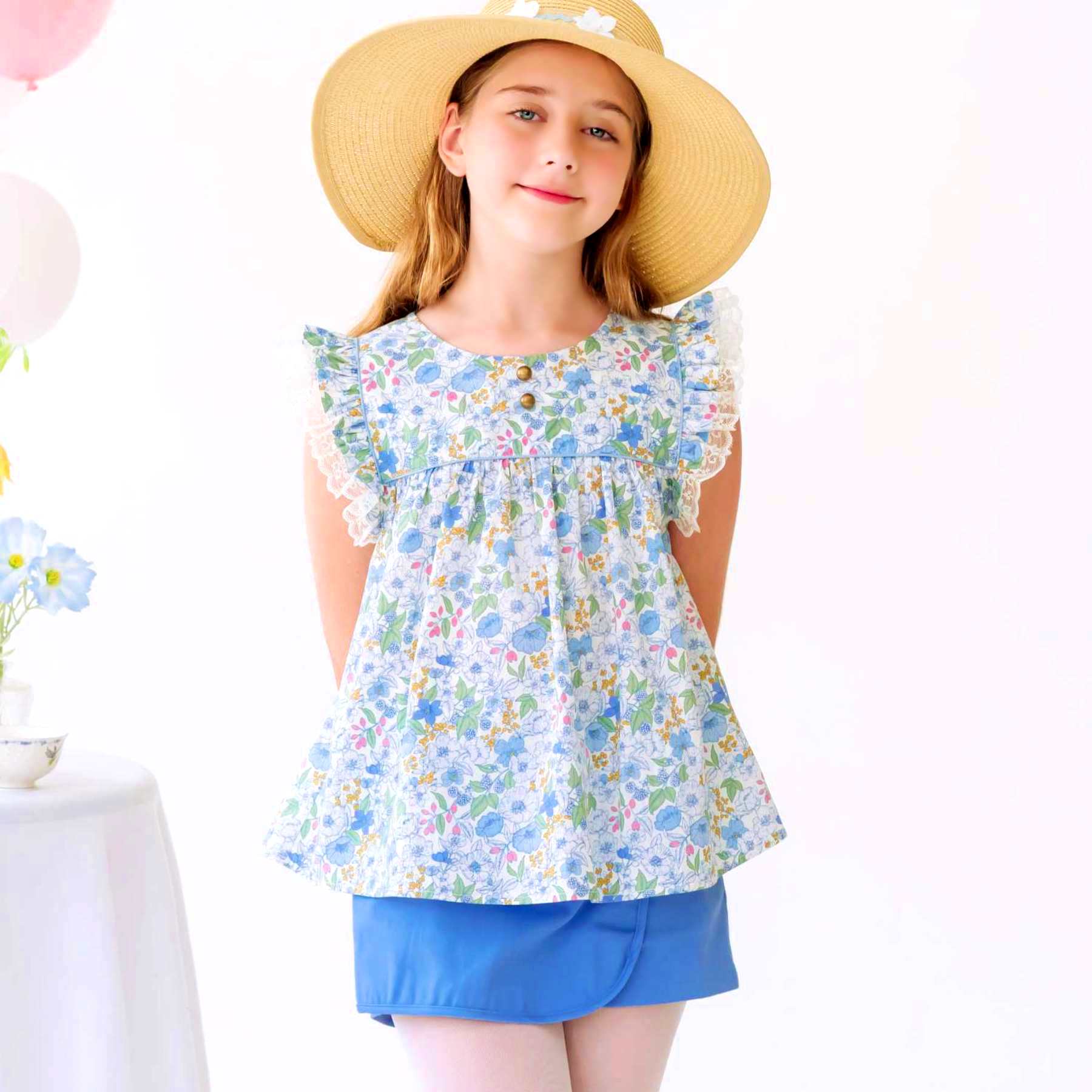 Photo portée de la blouse liberty fille fleurie bleu ciel avec manches courtes à volants bordés de fine dentelle beige. Col rond avec petits boutons ronds couleur cuivre et biais bleu ciel à paillettes. Fermeture boutons dans le dos. Tailles disponibles du 2 au 16 ans. Blouse portée avec la jupe short Jade bleu ciel. Cette blouse taille normalement.