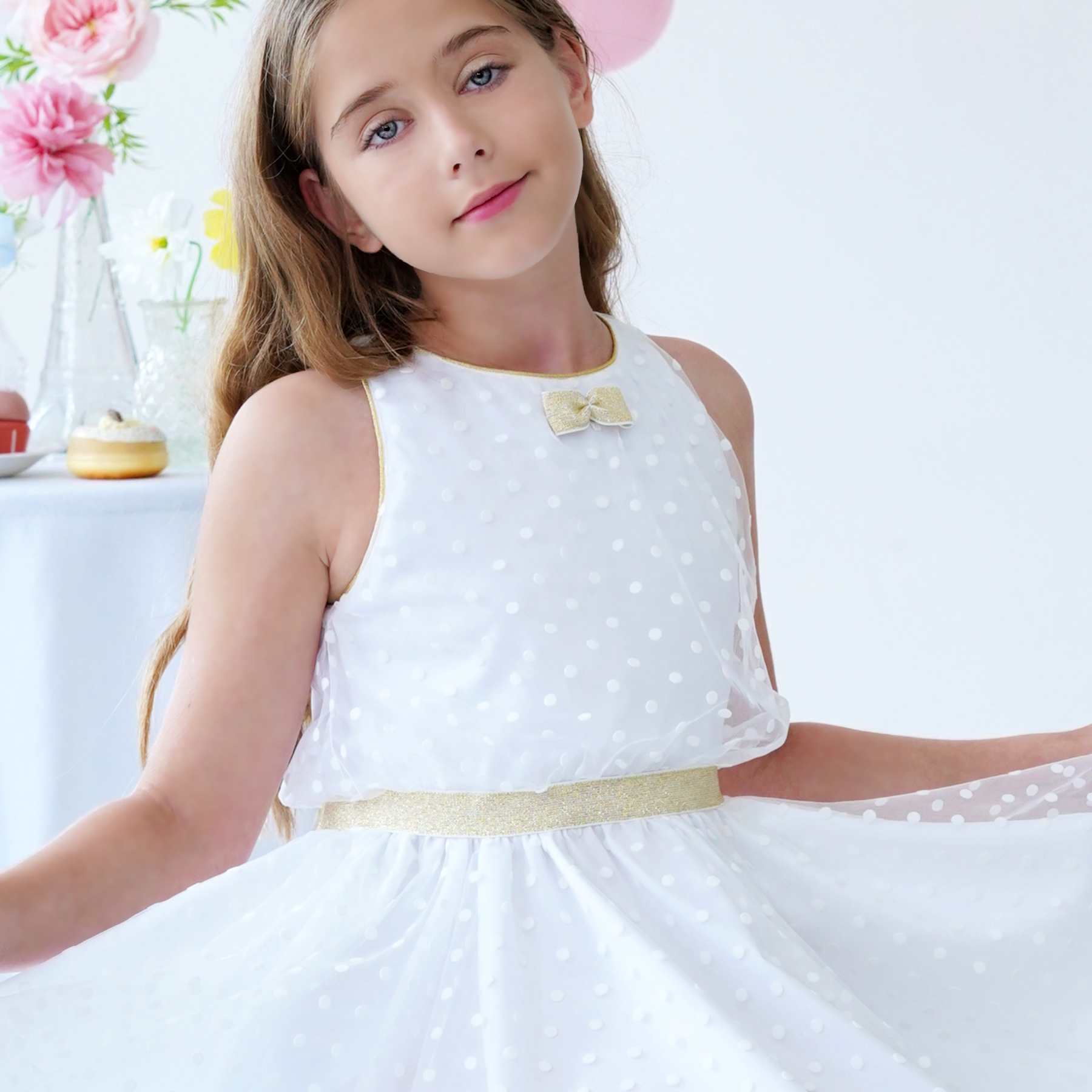 Gros plan haut de la robe de cérémonie fille blanche et or en tulle. Robe qui tourne coupe patineuse avec emmanchures américaines. Coupe trapèze avec taille cintrée dorée. Robe en tulle acrylique légère et douce doublé de coton confortable. Idéale pour les demoiselles d'honneur, les communiantes et tout évènements religieux. Fermeture dans le dos avec fermeture éclair et petits boutons ronds doré. Tailles disponibles du 2 au 16 ans.