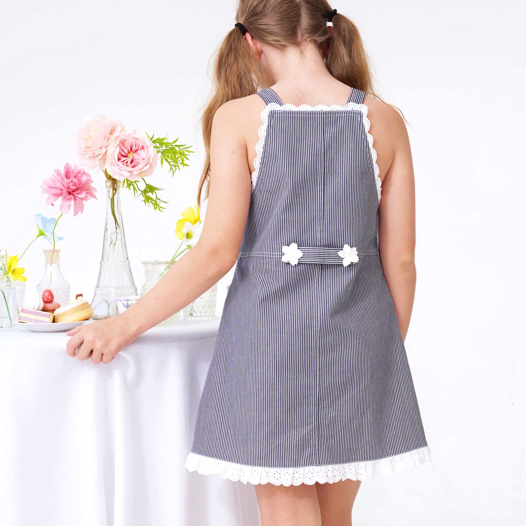 Dos de la robe salopette fille en jean rayé bleu marine avec broderie anglaise blanche Passepoil dans le dos avec deux fleurs blanches décoratives. Idéale en robe d'été pour l'école, les sorties en famille, la plage et les mariages champêtres. Disponible de la taille 2 à 16 ans, cette robe taille normalement. Optez pour la taille supérieure si votre enfant est entre deux tailles ou de morphologie ronde.