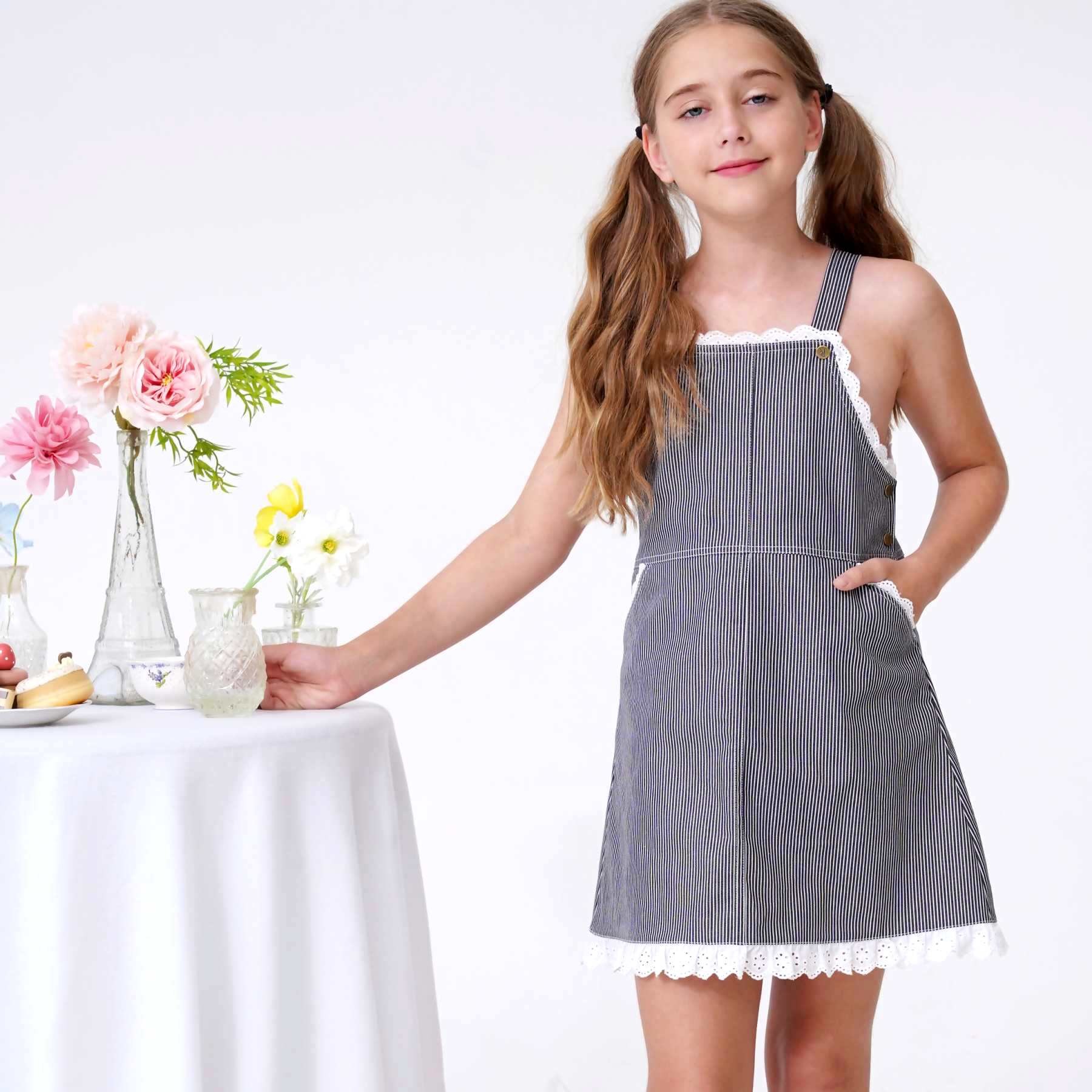 robe salopette fille en jean rayé bleu marine avec broderie anglaise blanche en haut, poches et bas de la robe. Bretelles ajustables. Idéale en robe d'été pour l'école, les sorties en famille, la plage et les mariages champêtres. Disponible de la taille 2 à 16 ans, cette robe taille normalement. Optez pour la taille supérieure si votre enfant est entre deux tailles ou de morphologie ronde.