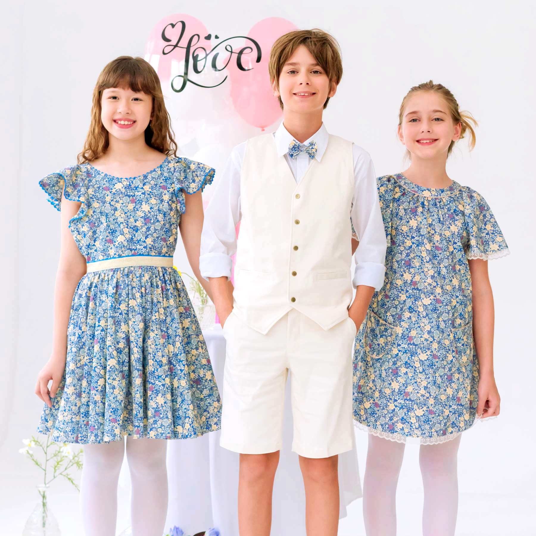 Photo portée tenues de cérémonie enfant avec bermuda de costume beige garçon Jimini à la coupe chino. Idéal porté avec le gilet de costume beige assorti Sainclair, chemise blanche col pointu Voltaire avec noeud papillon assorti aux robes de demoiselles d'honneur liberty.